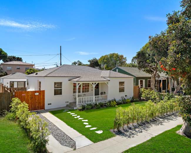 The image shows a small white house with a front porch, surrounded by a well-maintained lawn and flower beds, with a clear blue sky above and several trees in the vicinity.