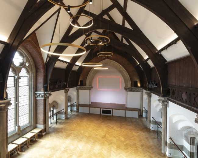 The image shows a spacious hall with high wooden beams, large windows, and an empty stage at one end.