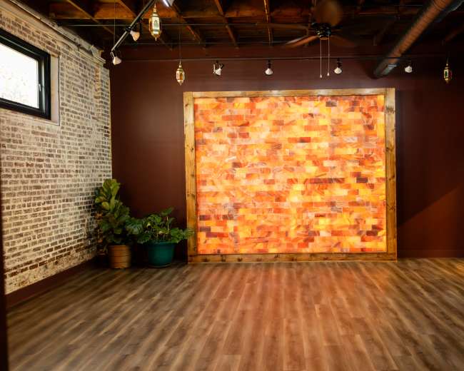 The image shows a spacious room with wooden flooring, a textured brick wall, and a large, colorful salt wall feature framed by wood.