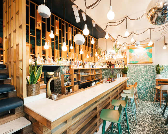 The image shows a modern bar with a wooden counter, colorful wall tiles, and a variety of liquor bottles displayed behind the bar.