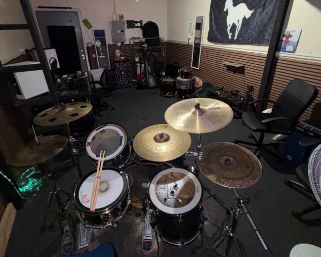 The image shows a home recording studio with a drum set in the foreground and various music equipment and chairs arranged around the room.