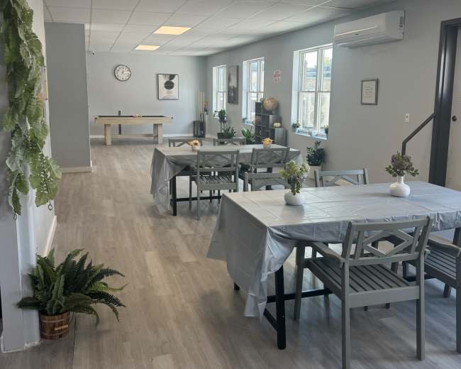 The image shows a spacious, well-lit interior with several tables set for dining, plants in pots, and a game area in the background.