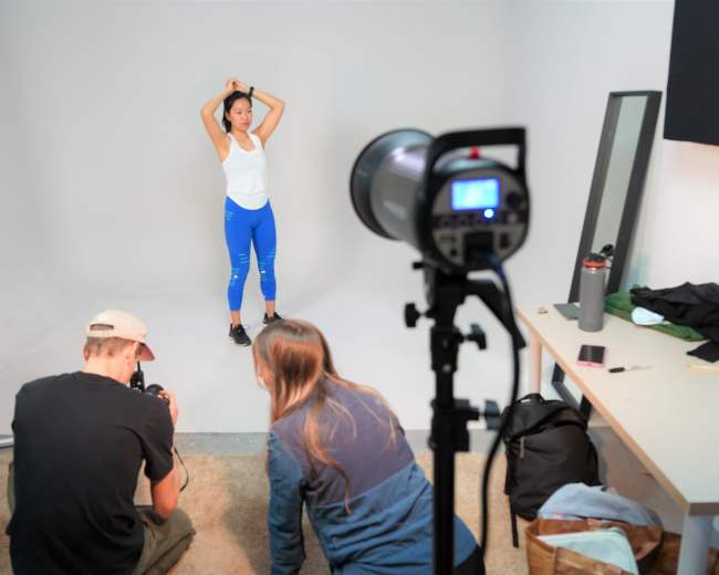 A model poses in a studio while two photographers set up equipment and capture images.