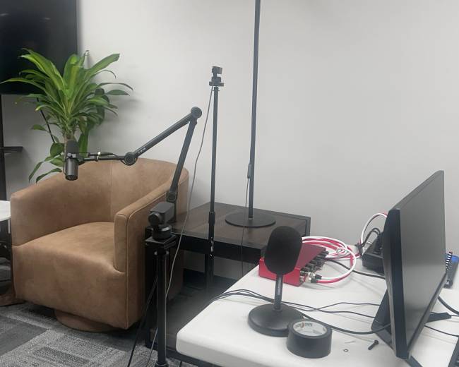 The image shows a small recording setup featuring a microphone on a stand, a computer monitor, and a brown chair against a plain wall with a plant in the background.