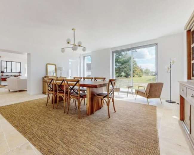 A spacious dining area features a wooden table surrounded by chairs, with large windows offering a view of the outdoors.