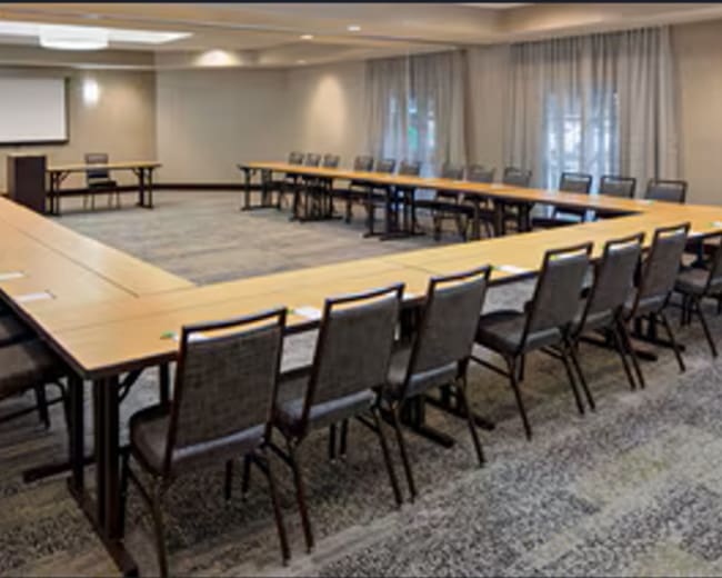 The image shows a conference room arranged with a large U-shaped table surrounded by chairs, with a projector screen at one end.