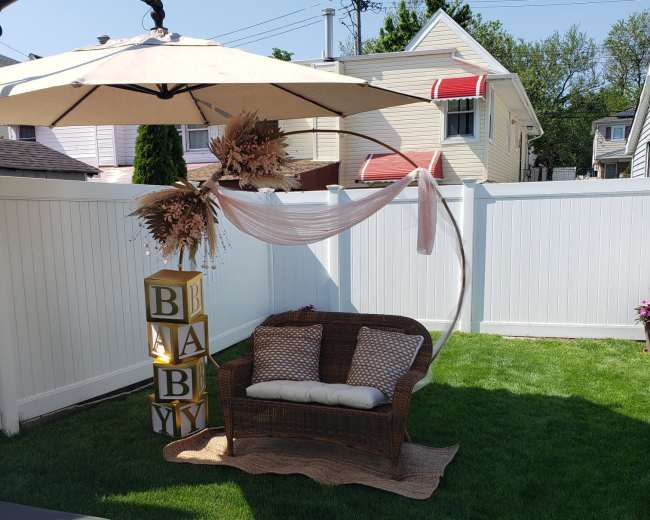 A wicker loveseat is positioned under a large umbrella, accompanied by decorative blocks spelling "BABY" and surrounded by a neatly maintained grass lawn and white fence.