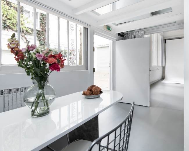 A bright, open space features a white table with a vase of flowers and a plate of croissants, surrounded by minimalist decor and natural light.