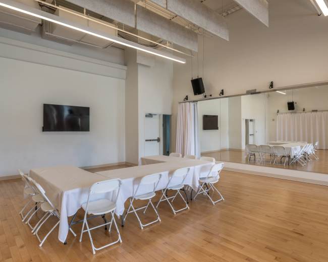 A spacious, well-lit meeting room features a long table with chairs arranged around it, a large wall-mounted TV, and a mirrored wall.