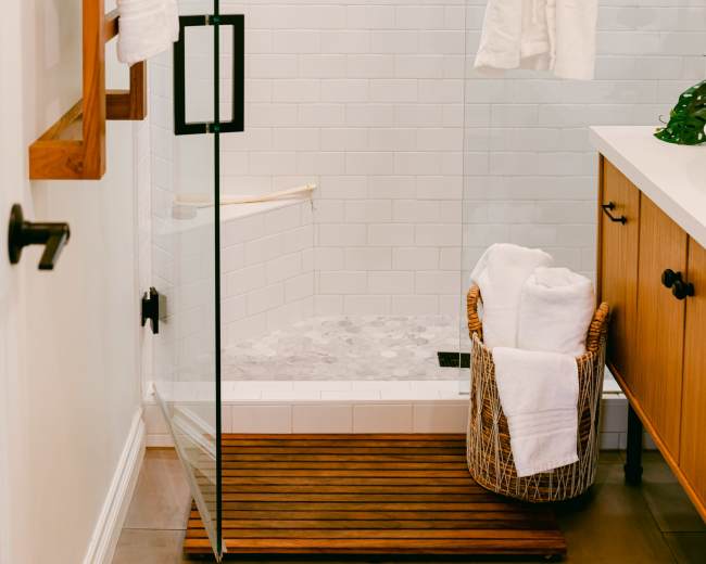 A glass shower enclosed by a wooden mat features a hanging white robe, with folded towels in a basket nearby and greenery visible outside a window.