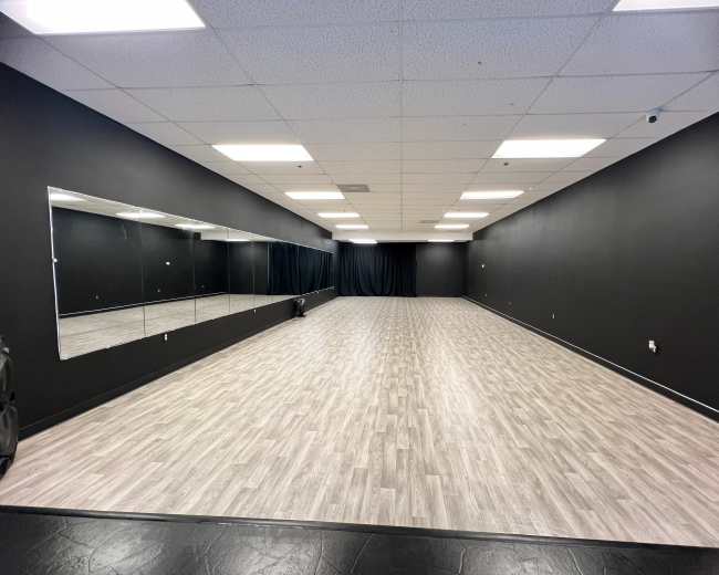 The image shows an empty dance studio with a polished wooden floor, black walls, and a large mirror along one side.