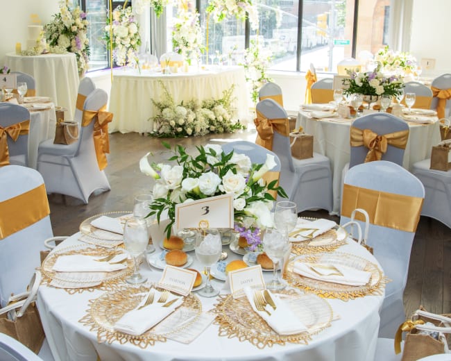 The image shows a elegantly decorated banquet room with round tables set for an event, featuring white and gold table settings and floral arrangements.