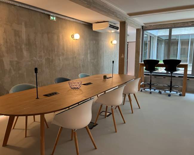 The image shows a modern conference room with a long wooden table surrounded by white chairs, equipped with microphones and power outlets, and a small bar area in the background.
