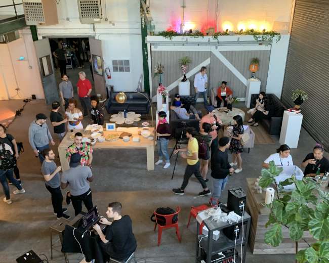 A lively gathering is taking place in a spacious, industrial-style venue with groups of people mingling around tables filled with food and drinks.