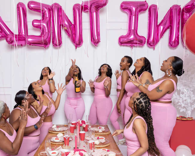 A group of women in matching pink outfits celebrates with pink balloons and decorations at a festive table filled with food and drinks.