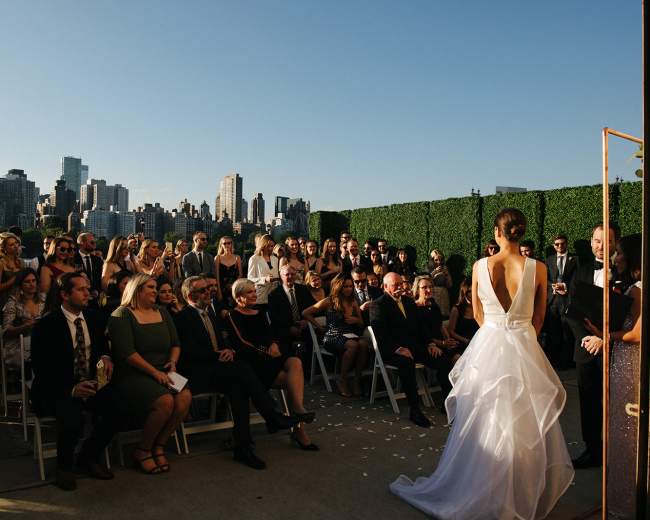 A bride wearing a white dress stands before a large audience at a wedding ceremony with a backdrop of a city skyline.
