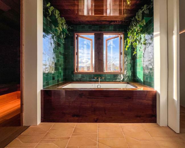 The image shows a modern bathroom with a green-tiled wall, a wooden bathtub, and two windows that provide natural light.