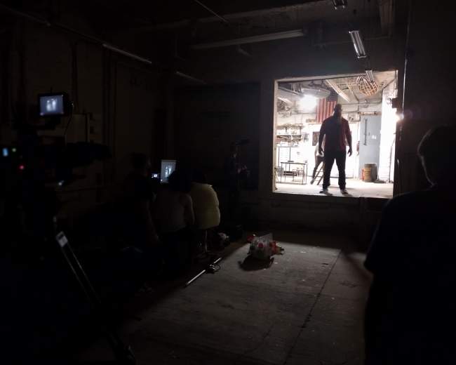 A dimly lit room features a figure silhouetted in a doorway with equipment and people seated in the foreground.