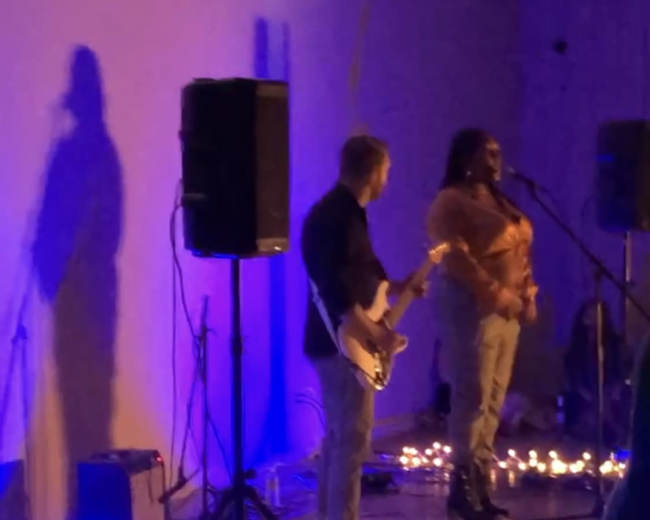 A singer performs on stage while a guitarist accompanies her, illuminated by blue and white lights with a backdrop of soft glowing decorations.