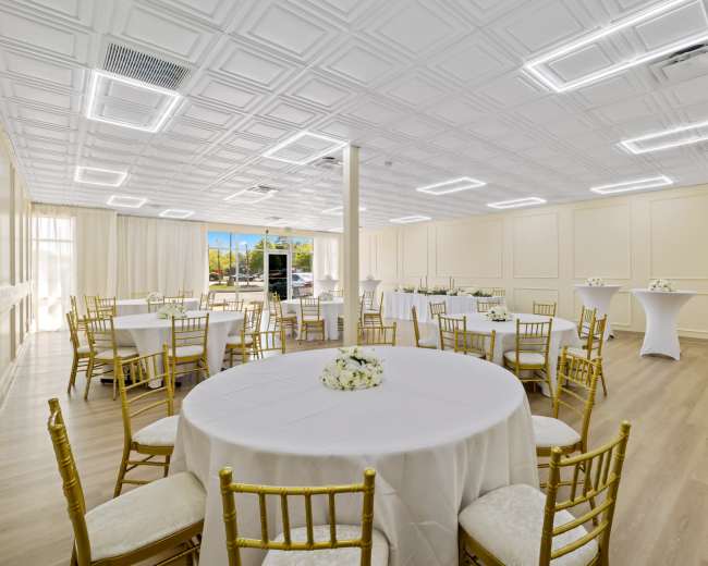 The image shows a spacious banquet room featuring several round tables with white tablecloths and gold chairs, along with a large window that lets in natural light.