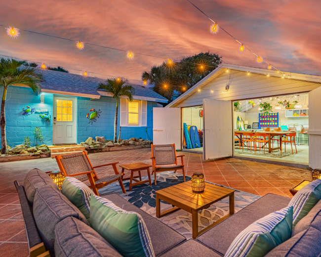 A cozy outdoor seating area with a fire pit is situated in front of a house, with string lights illuminating the scene against a colorful sunset sky.