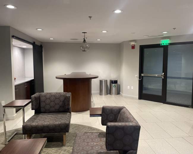 A modern lounge area with patterned chairs, a circular table, and a bar-like counter, next to a glass door and waste receptacles.