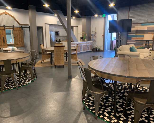 The image shows a modern communal space featuring multiple round wooden tables surrounded by metal chairs, a kitchenette visible in the background, and a cozy seating area.