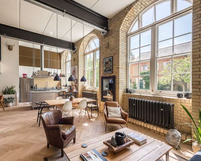 The image shows an open-concept living space featuring large windows, a modern kitchen area, a dining table, and a cozy seating arrangement with wooden flooring.