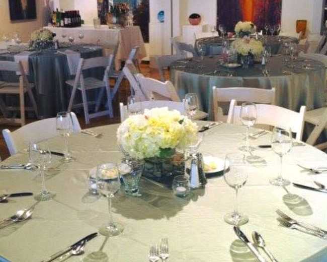 The image shows a neatly arranged dining area with round tables covered in light fabric, set with glassware and silverware, surrounded by modern art on the walls.
