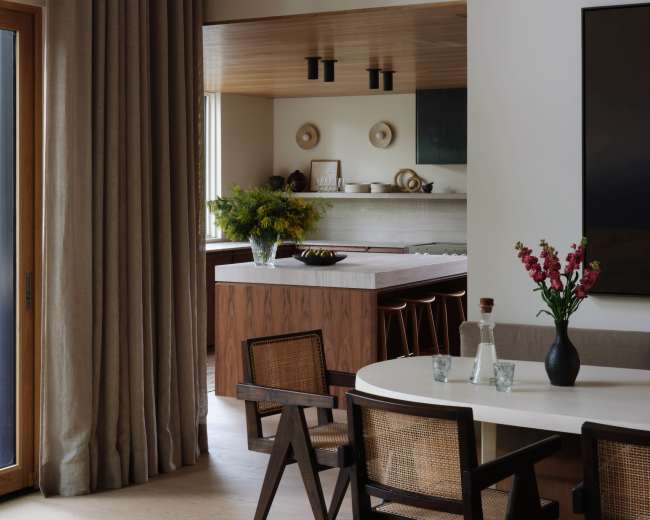 A modern interior features a dining area with a round table surrounded by wooden chairs, set against a backdrop of large windows and soft drapery.