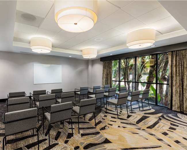 The image shows a meeting room arranged with chairs facing a blank whiteboard, surrounded by large windows with a view of outdoor greenery.
