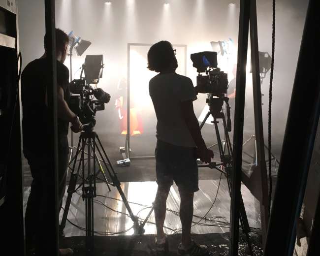 A film crew sets up cameras and lighting in a dimly lit studio, with smoke and backlighting creating a dramatic atmosphere.