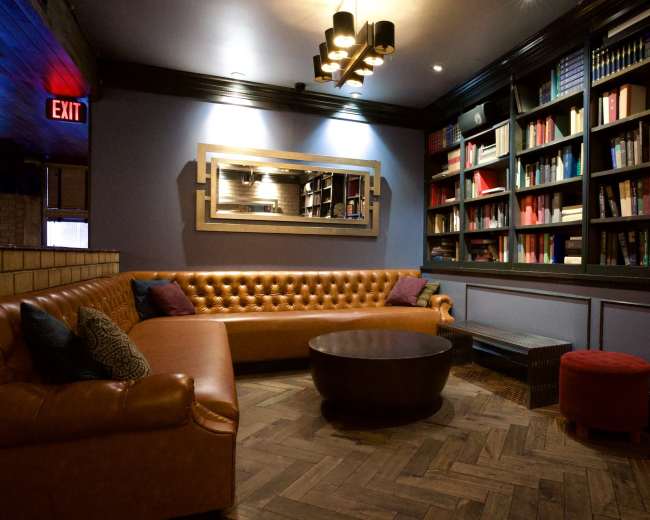 The image shows a cozy lounge area with a brown leather sofa, a round wooden coffee table, and a bookshelf filled with books, highlighted by soft lighting and a mirror on the wall.