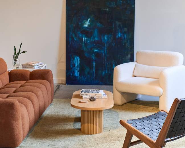 The image shows a modern living room with a brown sofa, a white armchair, a round wooden coffee table, and a textured area rug, set against a wall with an abstract blue painting.