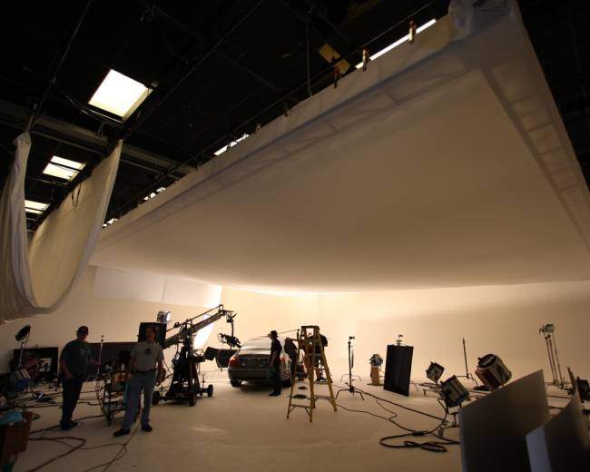 A film studio setup features a suspended white backdrop, lighting equipment, cameras, and crew members preparing for a shoot.