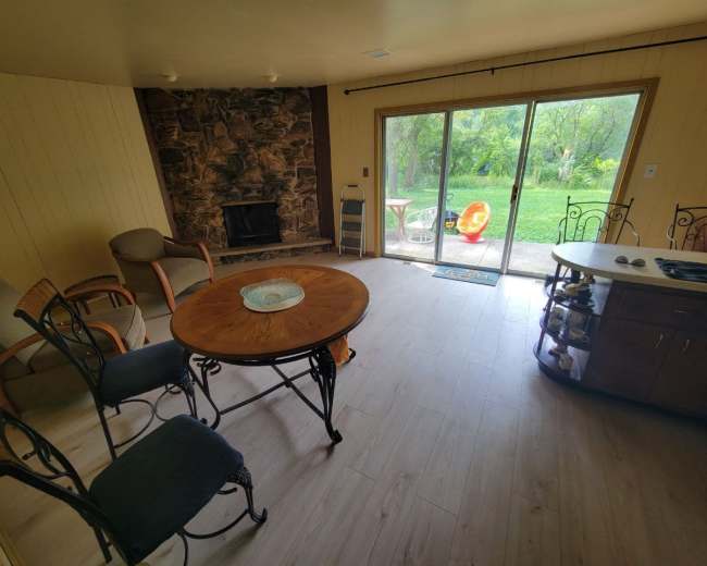 The image shows a cozy indoor living space with a round dining table and chairs, a stone fireplace, and large sliding glass doors leading to a green outdoor area.