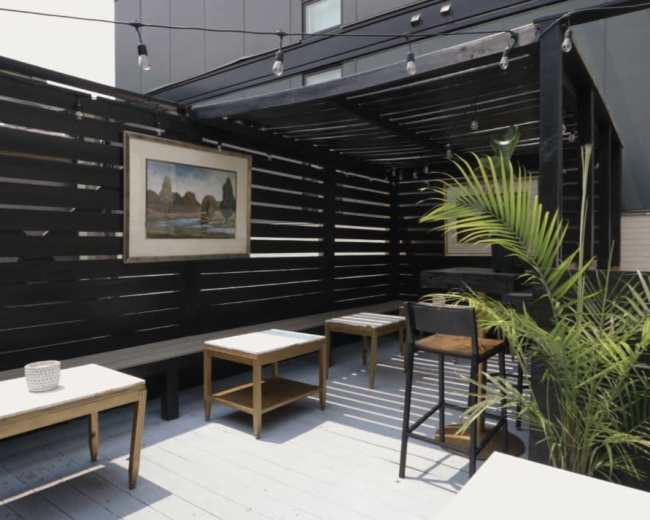 The image features a small outdoor seating area with wooden tables, a chair, a plant, and soft lighting, enclosed by black wooden slats.