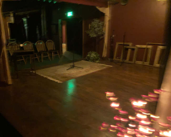 A dimly lit performance space features a wooden floor, a small rug, and several chairs arranged around a table.