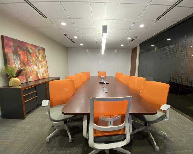A conference room features a long wooden table surrounded by orange chairs, with a modern art piece and a plant on the wall.