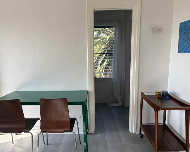 A green table with two wooden chairs is positioned in a brightly lit room next to an open doorway leading to another space with palm trees visible through the window.