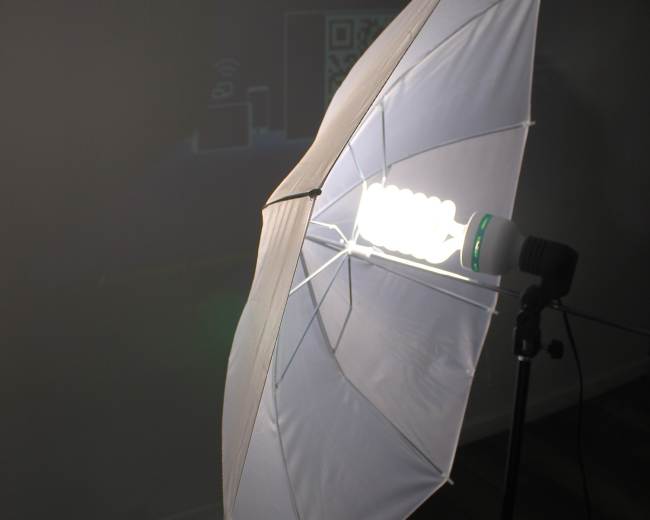 A photography umbrella softbox is mounted on a stand, with a large bulb visible inside, casting light onto a nearby wall.