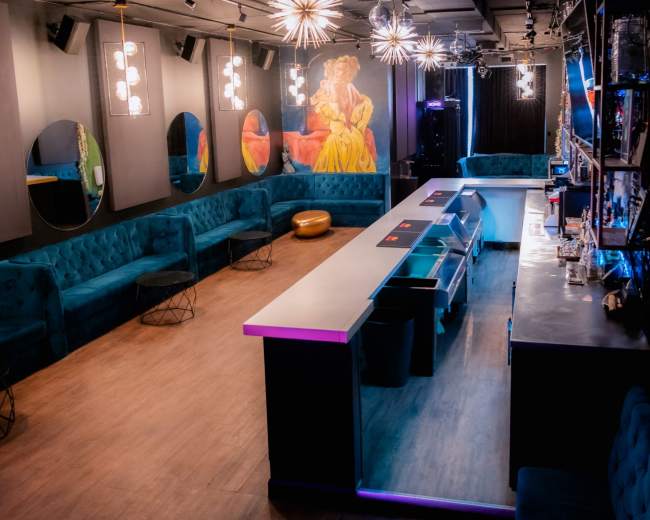 The image shows a stylish lounge area with plush teal seating, a long bar with illuminated edges, and artistic wall decor.