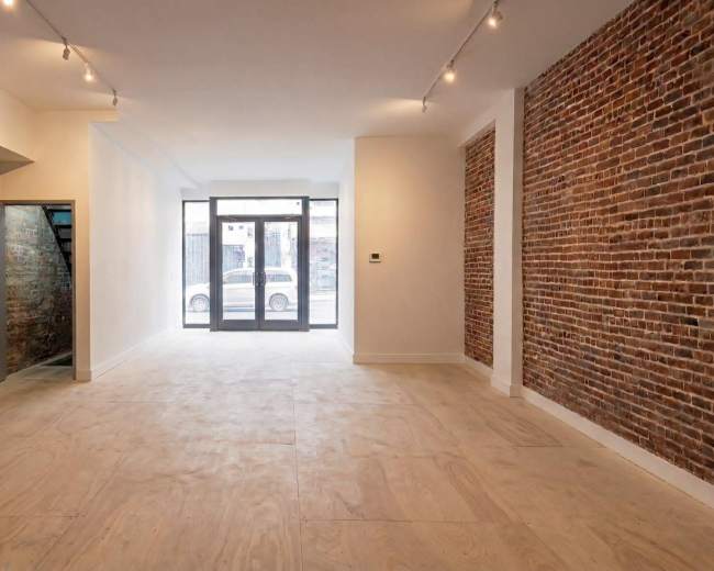 The image shows an empty interior space with exposed brick walls, wood flooring, and large windows facing the street.
