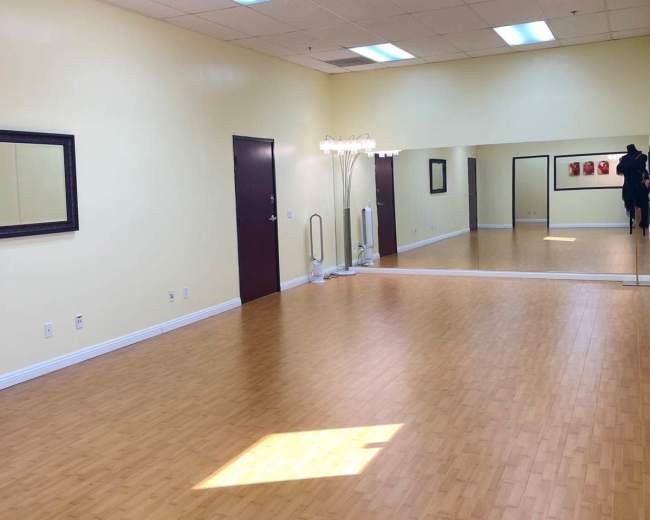 The image shows an empty dance studio with wooden flooring, mirrored walls, and soft yellow paint.