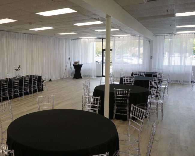 The image shows an empty event space with black round tables and transparent chairs arranged in a clean, modern layout, surrounded by white drapes and bright overhead lighting.