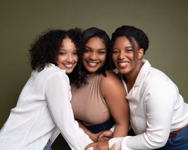 Three women stand closely together, smiling at the camera against a green background.