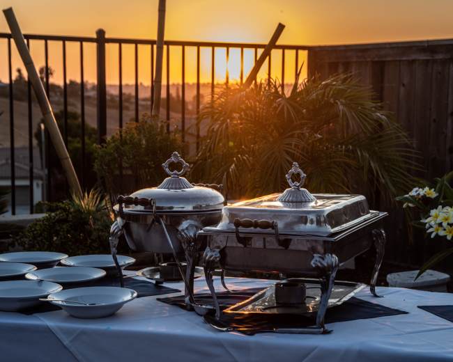 Two silver chafing dishes are set on a table with white plates, overlooking a sunset behind a fence and plants.