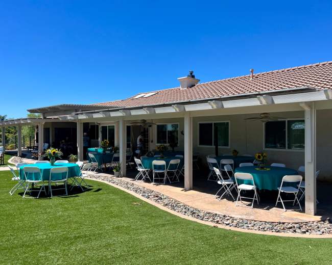 A backyard event setup features tables with teal tablecloths arranged on a green lawn next to a house with a tile roof.