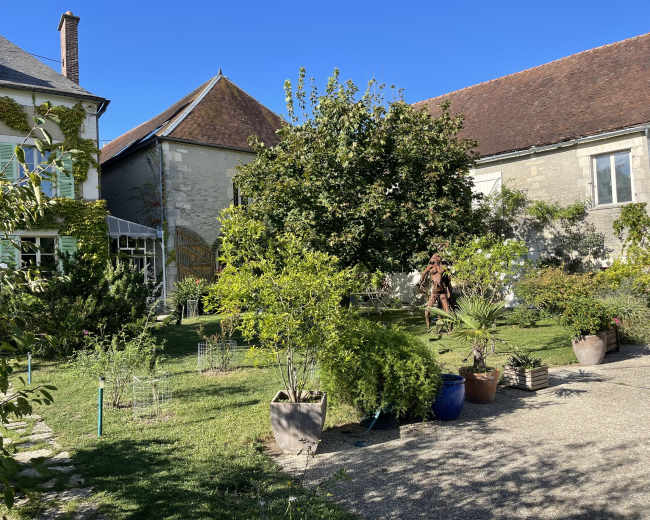 The image shows a serene garden scene featuring a variety of plants and trees surrounding two buildings with blue skies in the background.