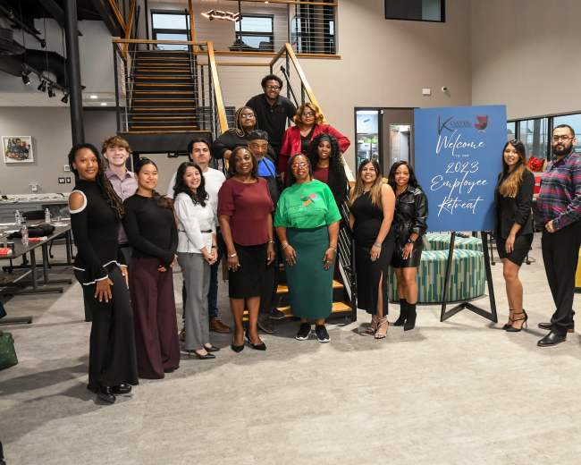 A group of diverse individuals stands together at an indoor venue, posing for a photo in front of a welcome sign for a 2023 employee retreat.